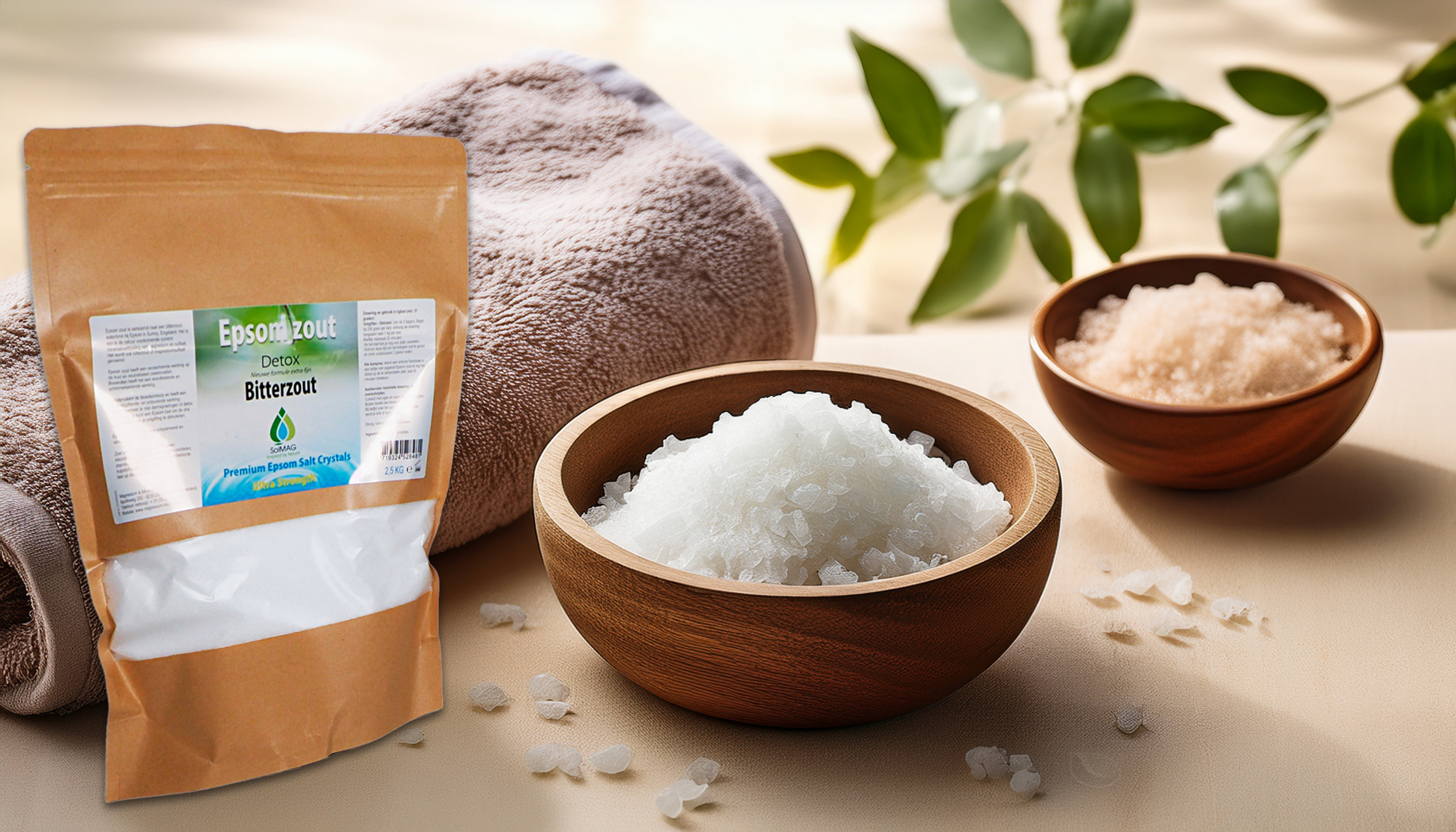 Verpackung von Bittersalz (Epsom-Salz) neben Holzschalen mit weißen und rosafarbenen Magnesiumsulfatkristallen, in einem Wellness-Umfeld mit Handtuch und grünen Blättern.